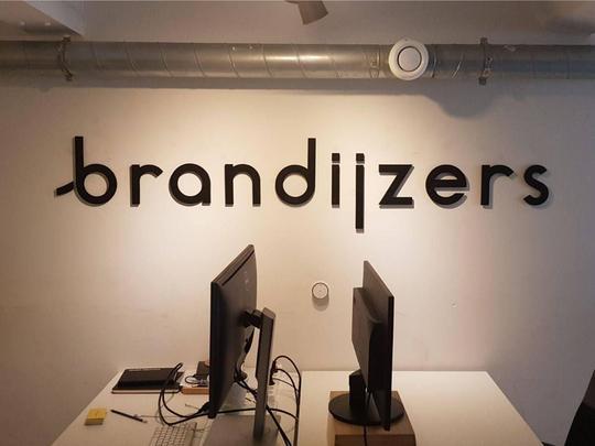 Zwart woordlogo brandijzers op een witte kantoorwand boven twee beeldschermen
